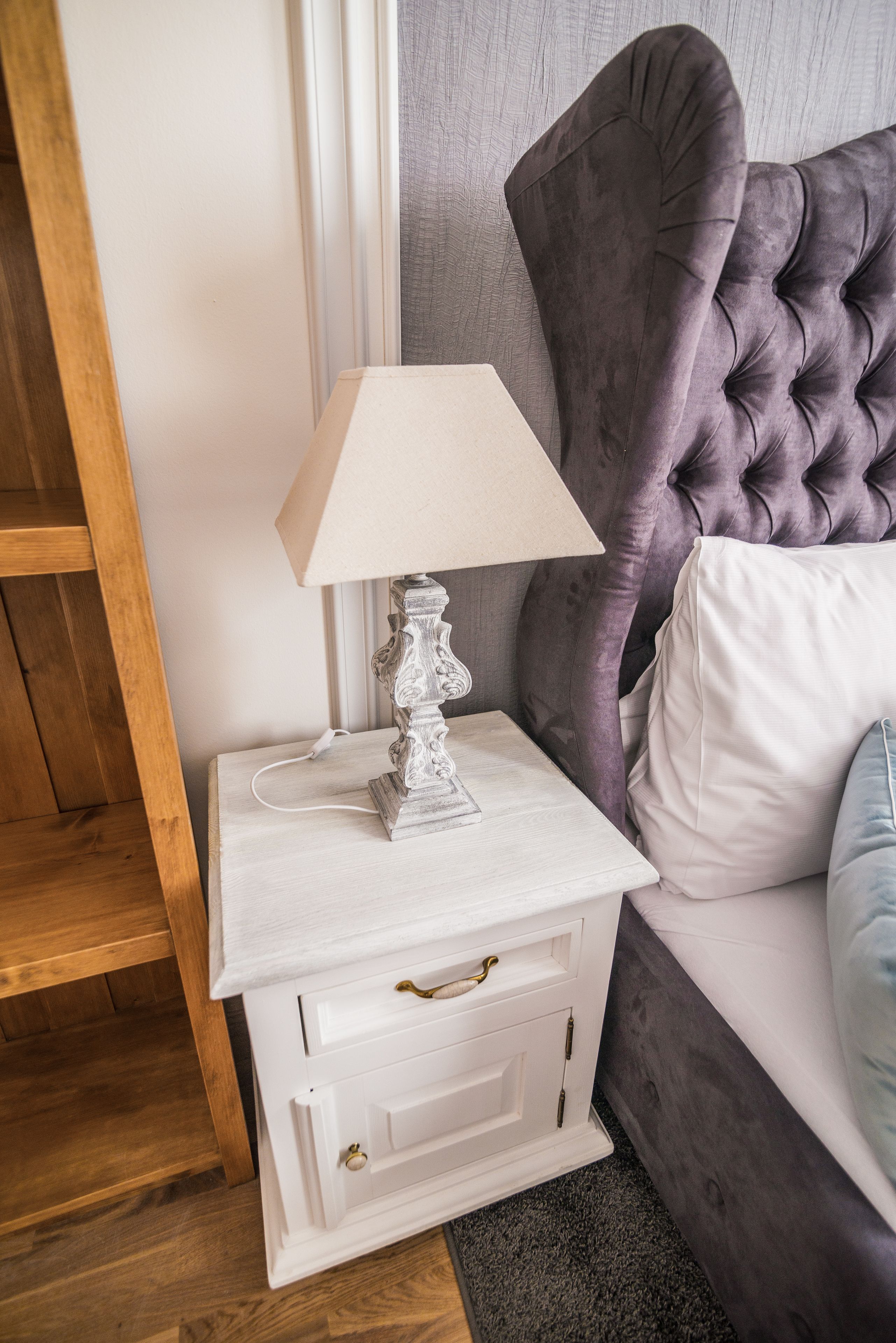 Une table de chevet blanche avec une lampe, une tête de lit recouverte d’un tissu violet et un oreiller blanc.