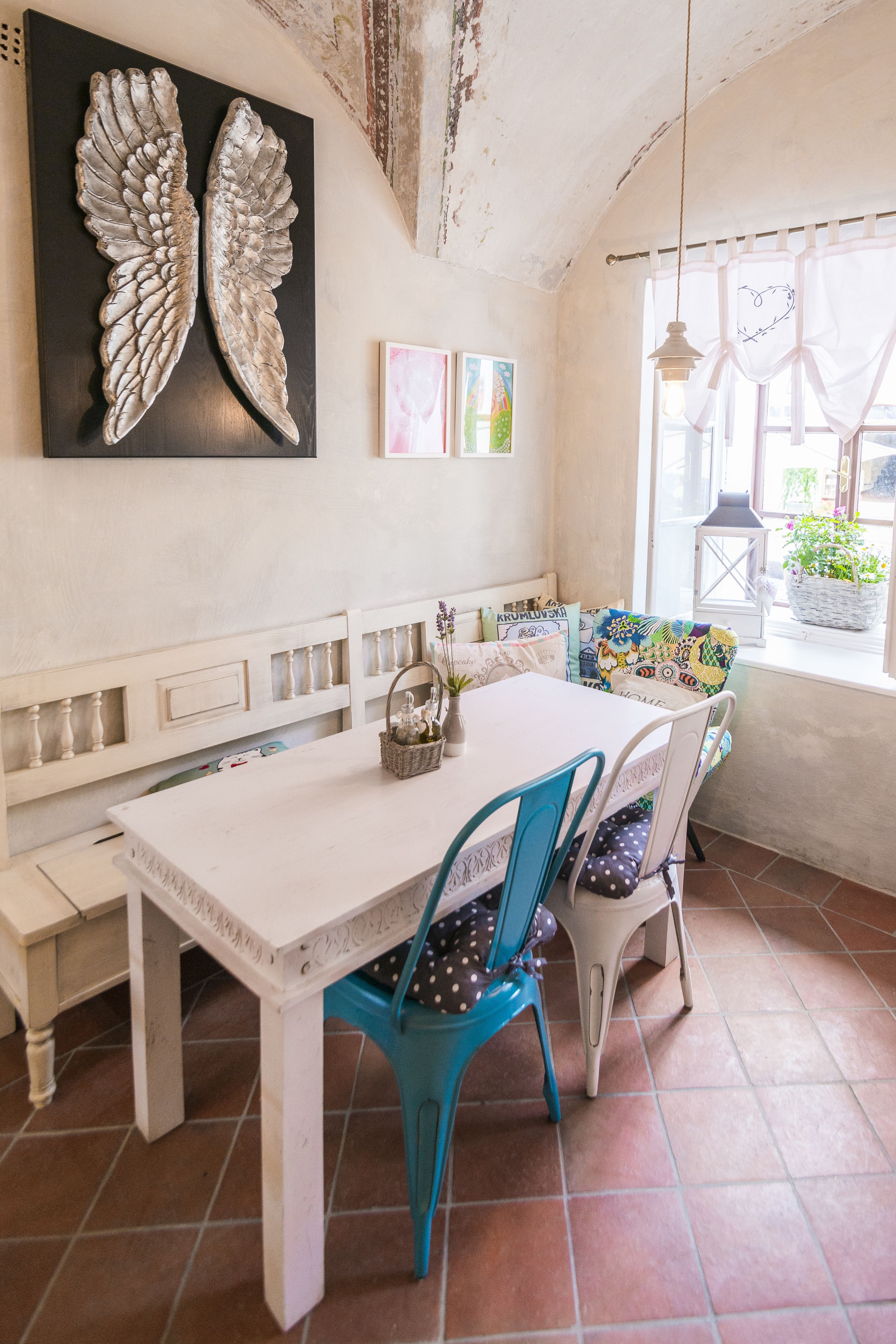 A dining area with a white table, blue chairs, and a bench. There is a decorative wall piece above the table and a window with curtains.