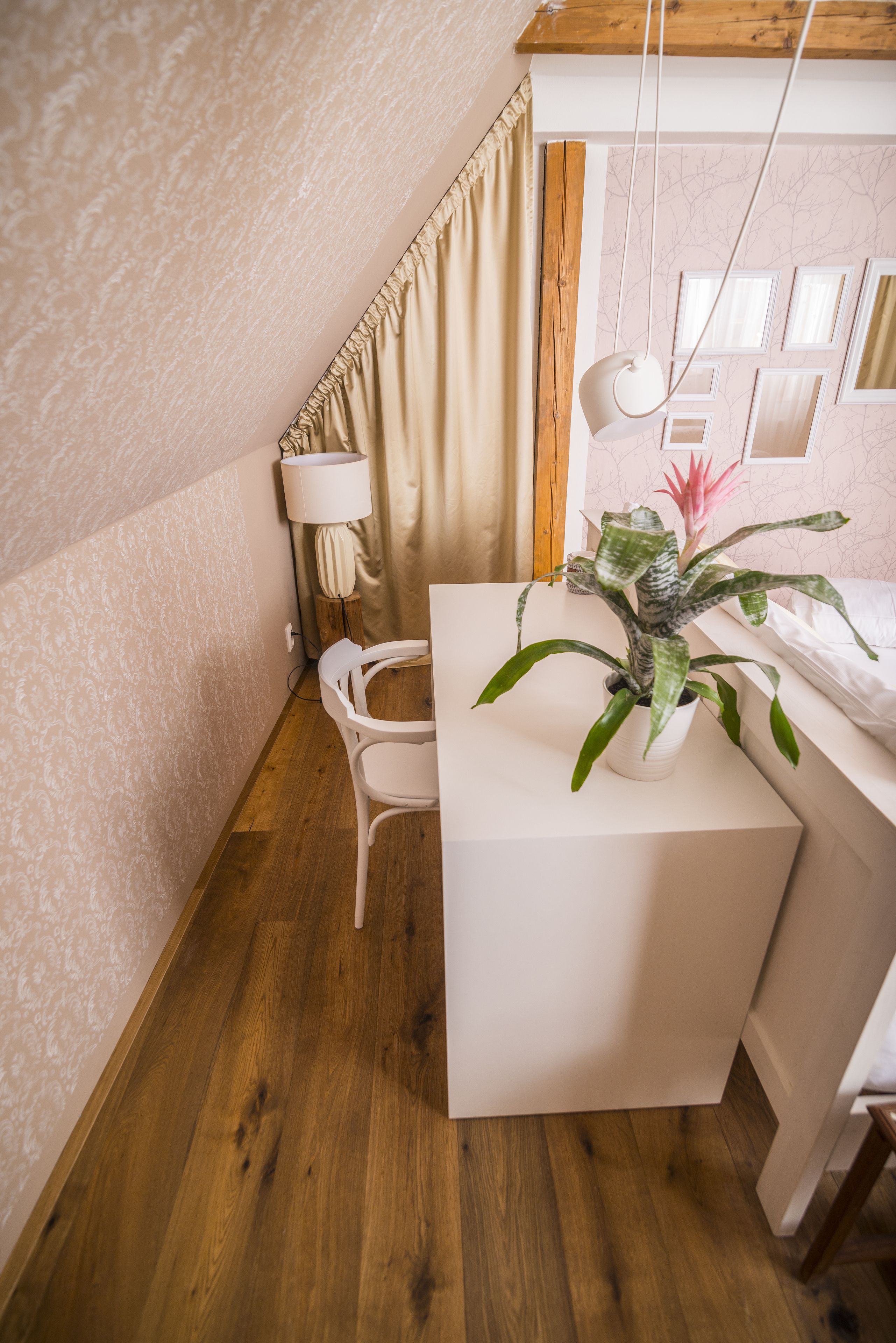 A small, cozy room with a wooden floor, a white desk, a chair, a potted plant, a lamp, and a window with curtains.