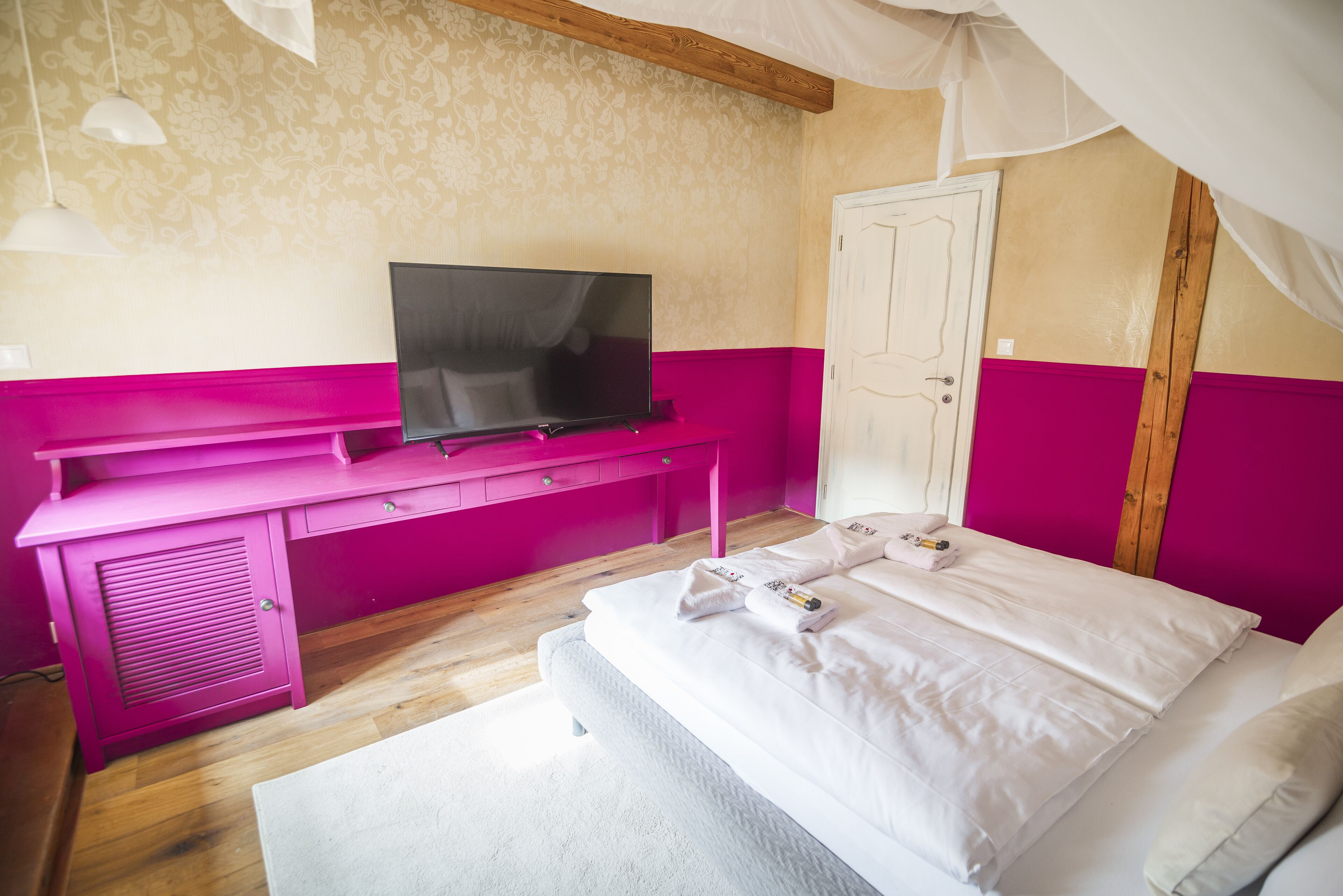 A bedroom with a bed, a TV on a pink desk, and a white door.