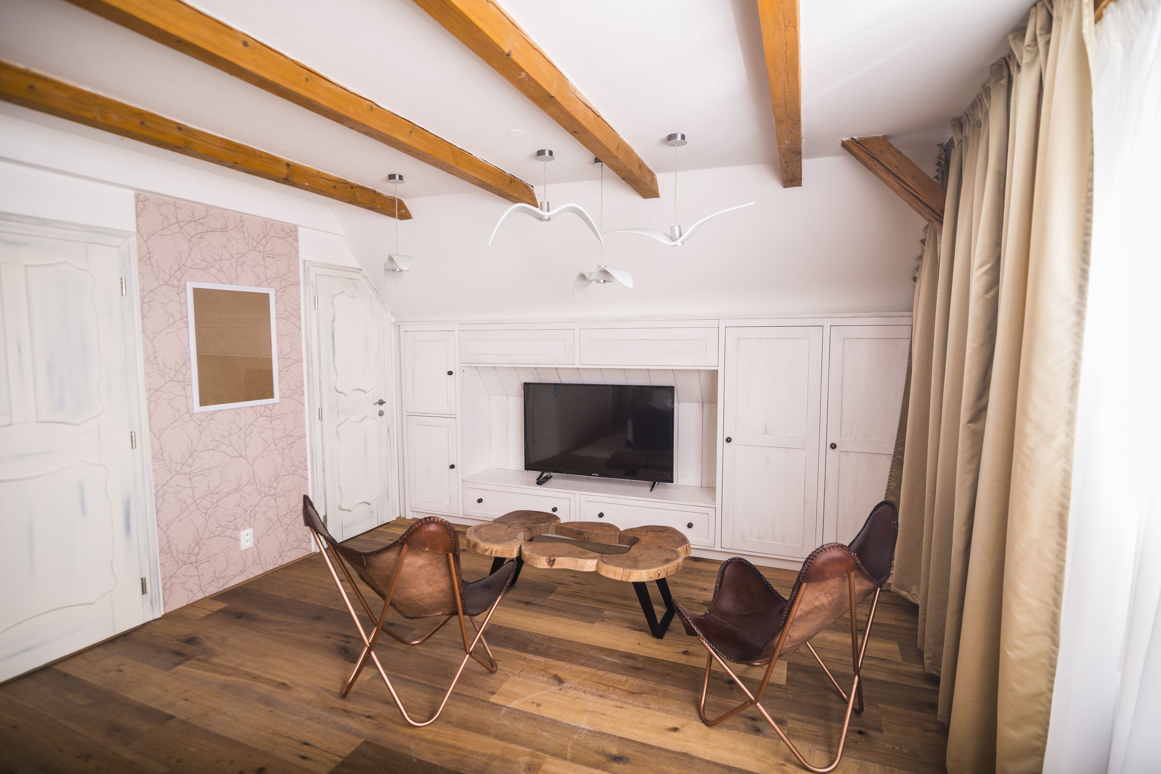 A spacious living room with a large flat-screen TV, wooden ceiling beams, and a fireplace.