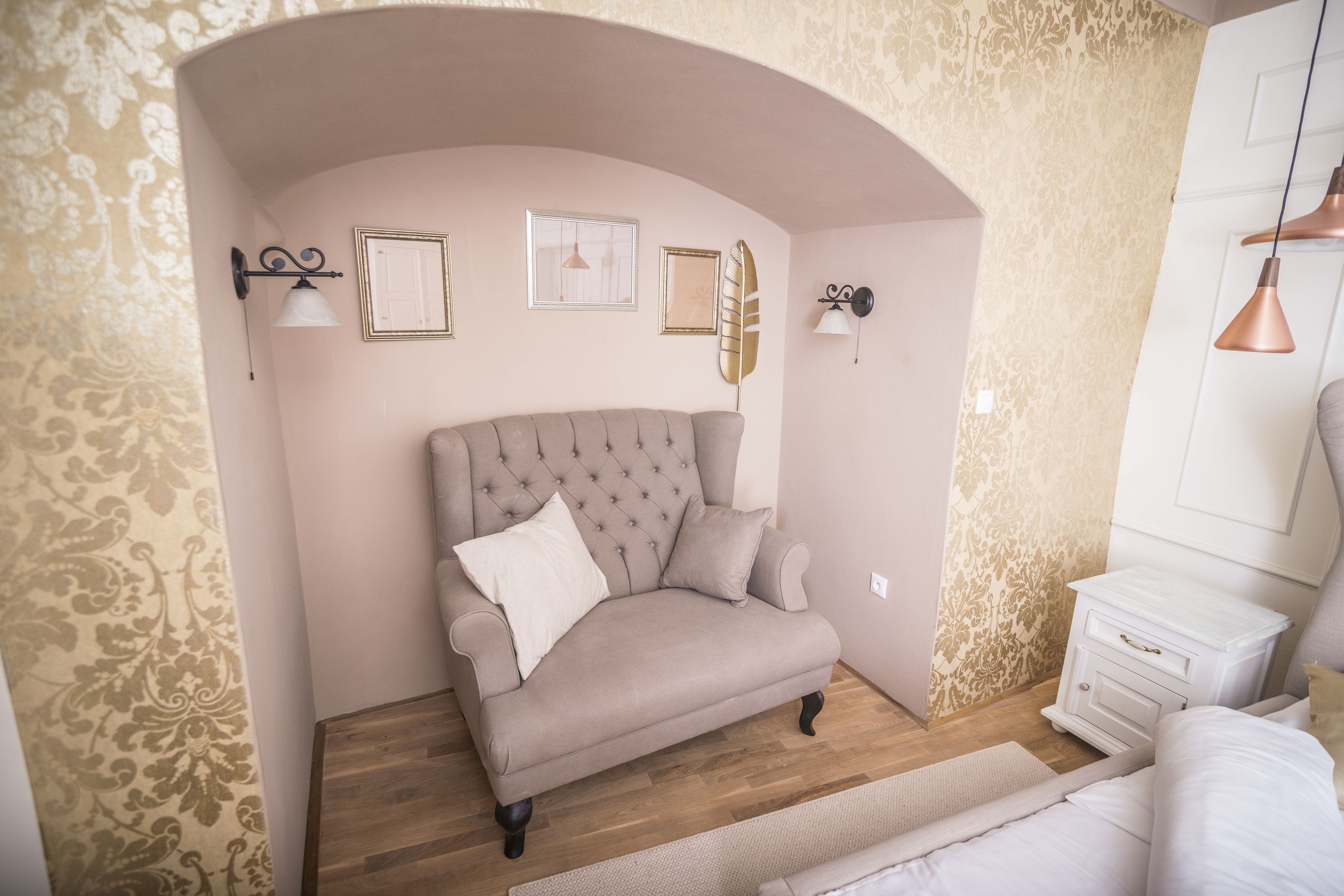 A bedroom with a tufted sofa, a white bedside table, and a decorative wall with framed artwork.