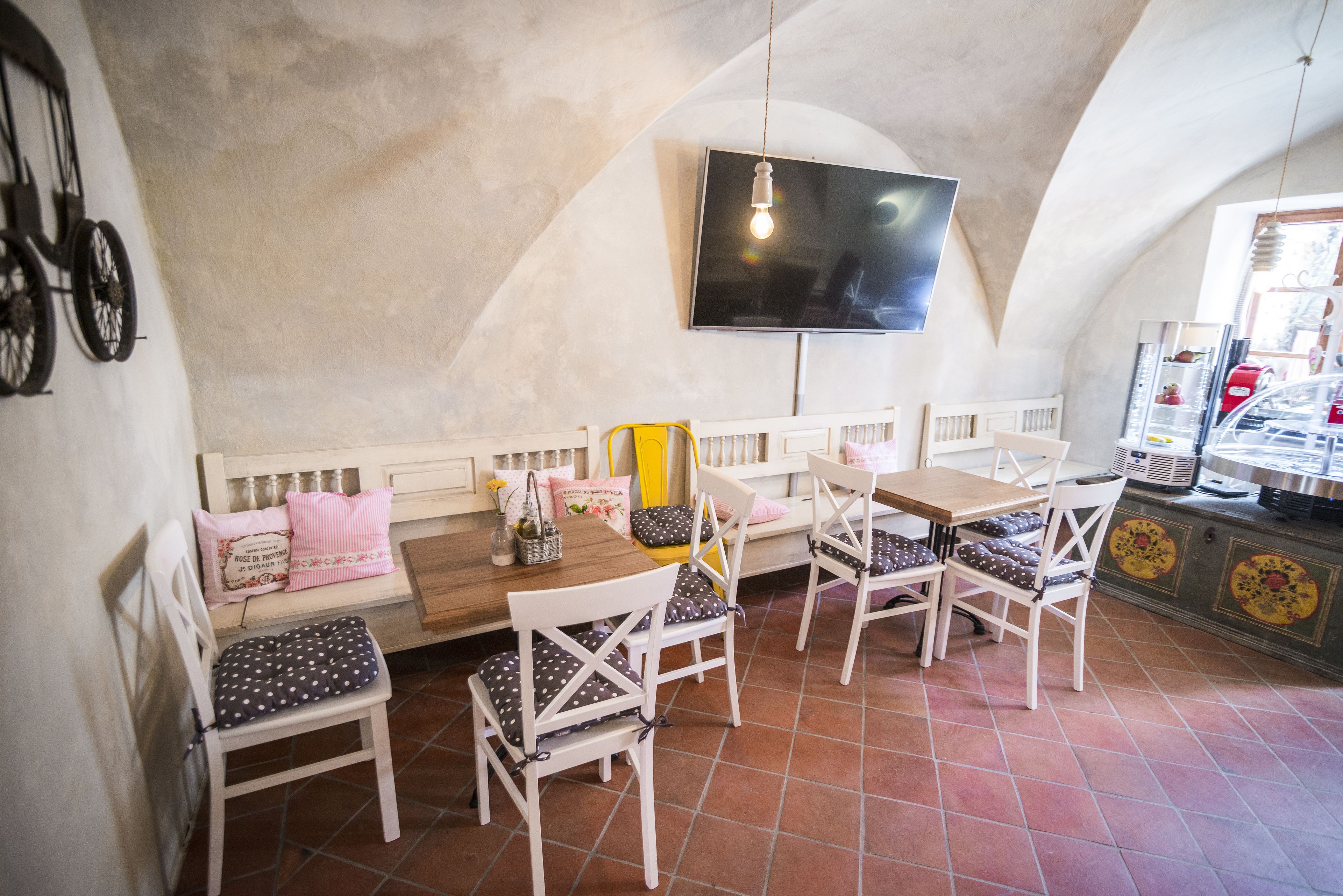 Un espace repas avec un long banc, des chaises blanches et un téléviseur fixé au mur.
