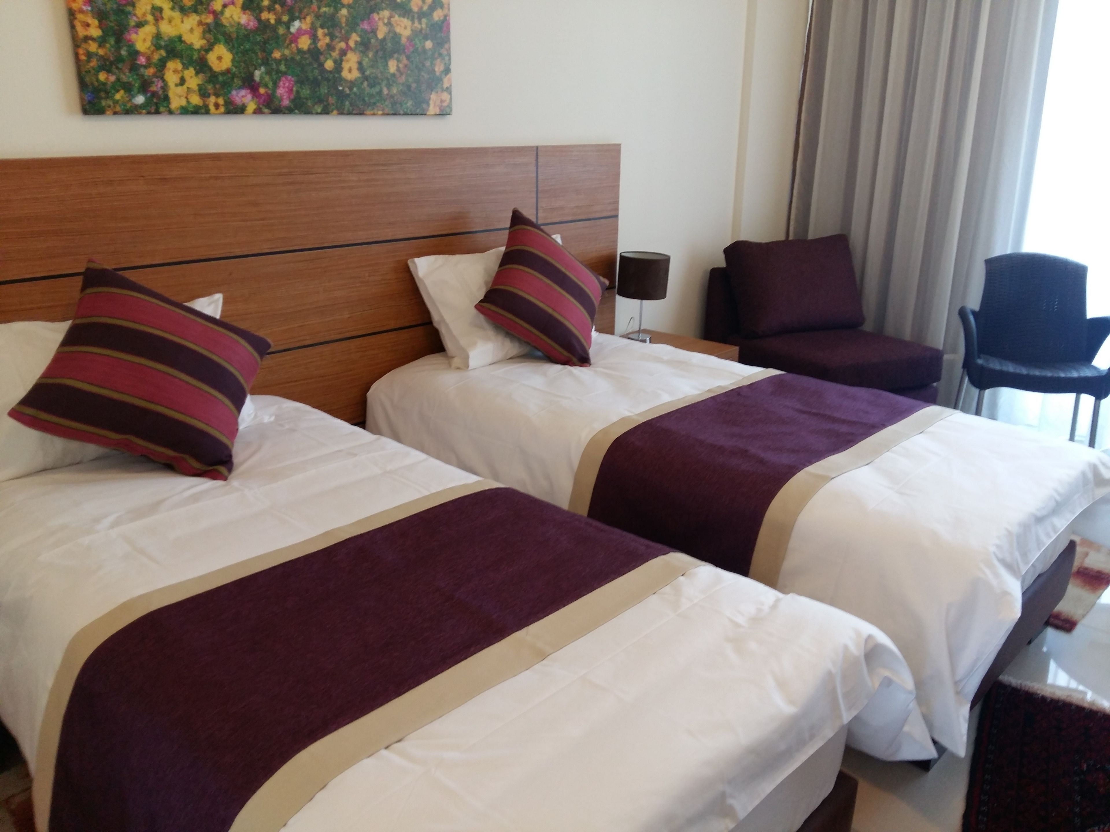 Two beds with white linens and purple accents, a wooden headboard, a chair, and a painting on the wall.