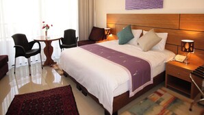 Een hotelkamer met een groot bed, een nachtkastje met een lamp, een stoel, een kleine tafel met een bloemenvaas, en een schilderij aan de muur.