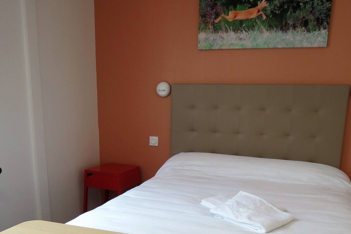 A bedroom with a bed, a red chair, a small red table, and a painting of a deer on the wall.