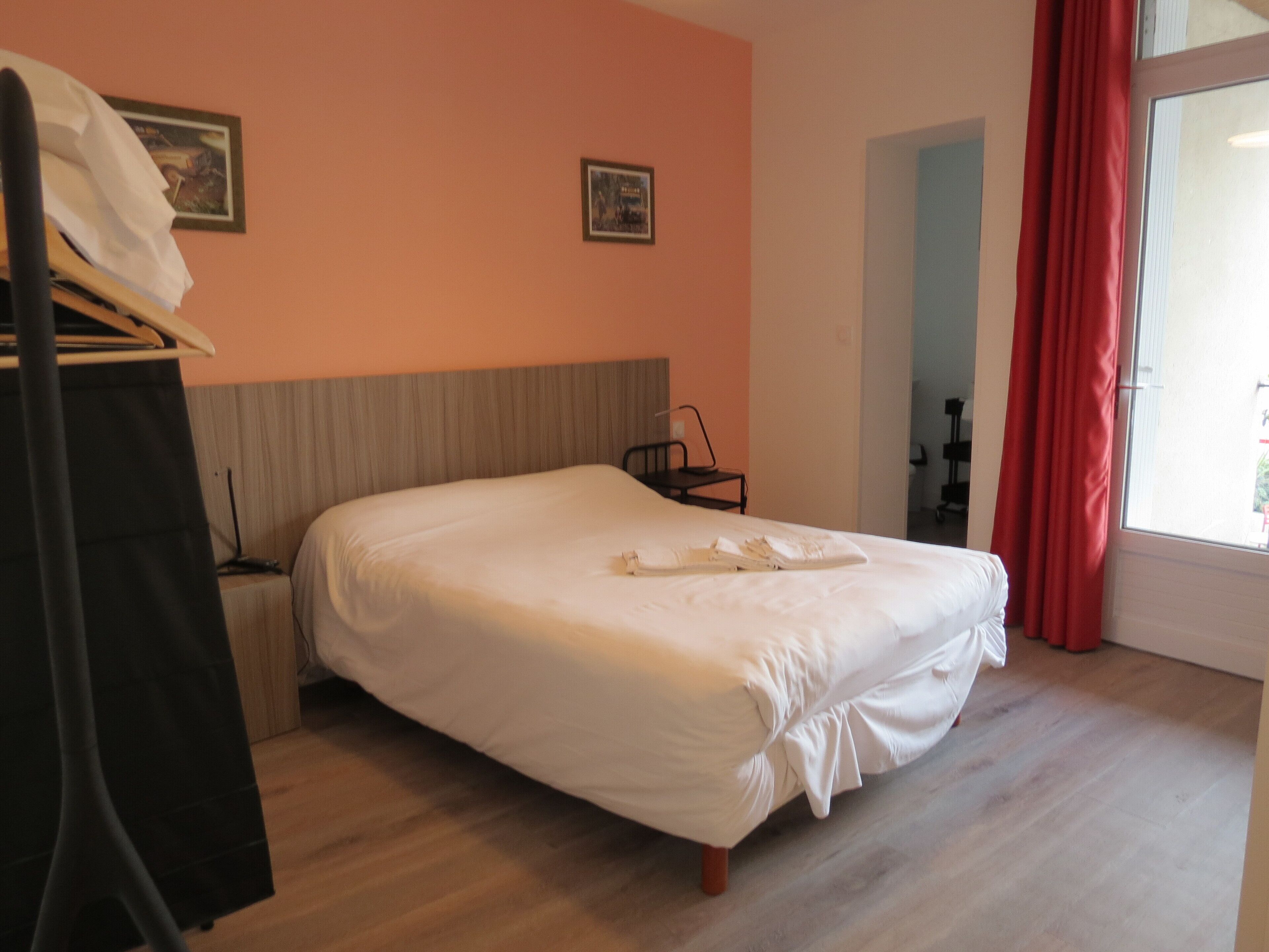 Una cama bien hecha en una habitación con suelo de madera, cabecera y ventana con cortinas rojas.