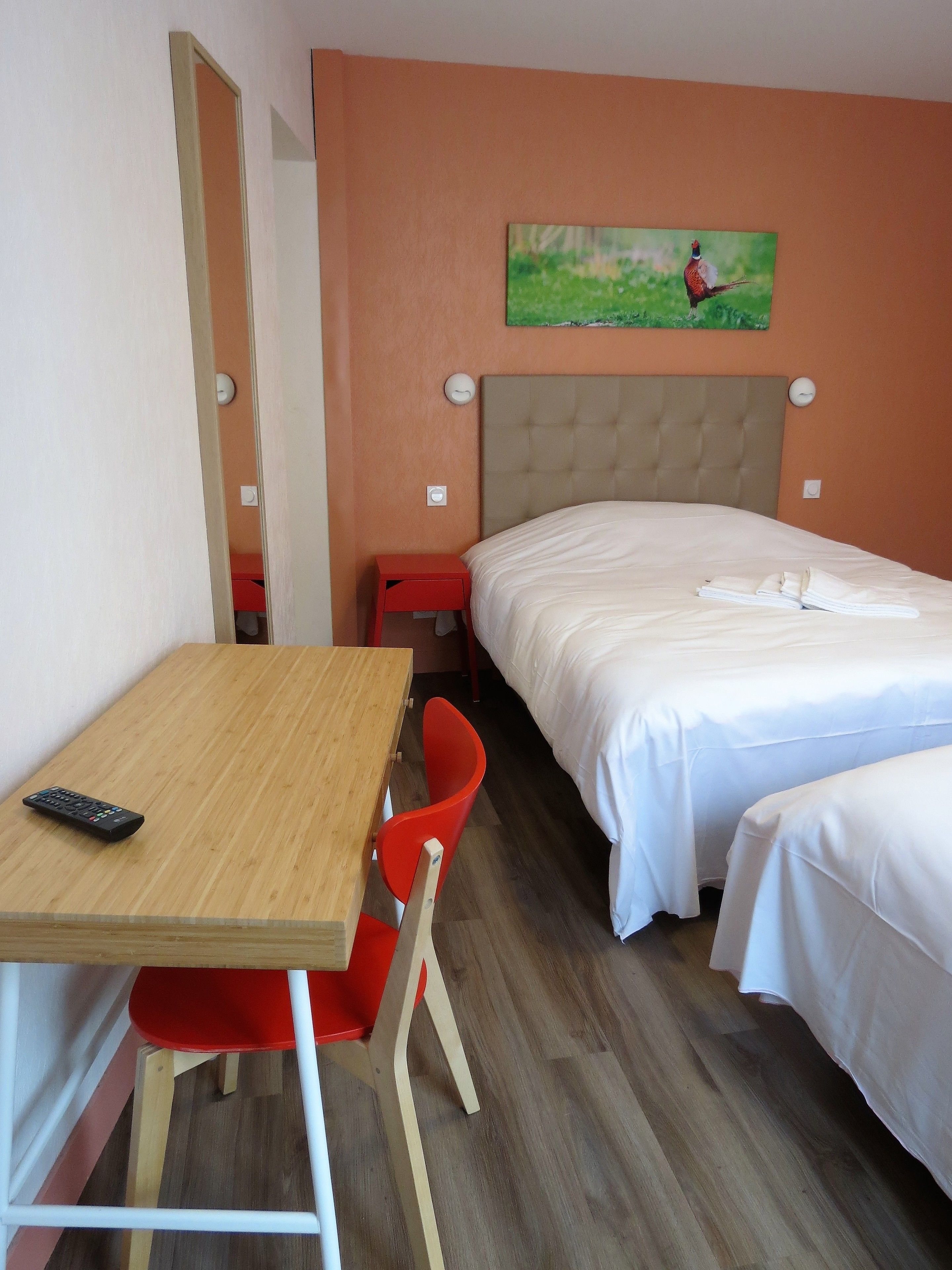 A hotel room with a wooden desk, a red chair, a bed with white bedding, and a painting on the wall.