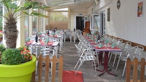 A covered outdoor dining area with tables and chairs, a large potted plant, and a wooden floor. - Hotel Le Faisan (Saint-Avertin)