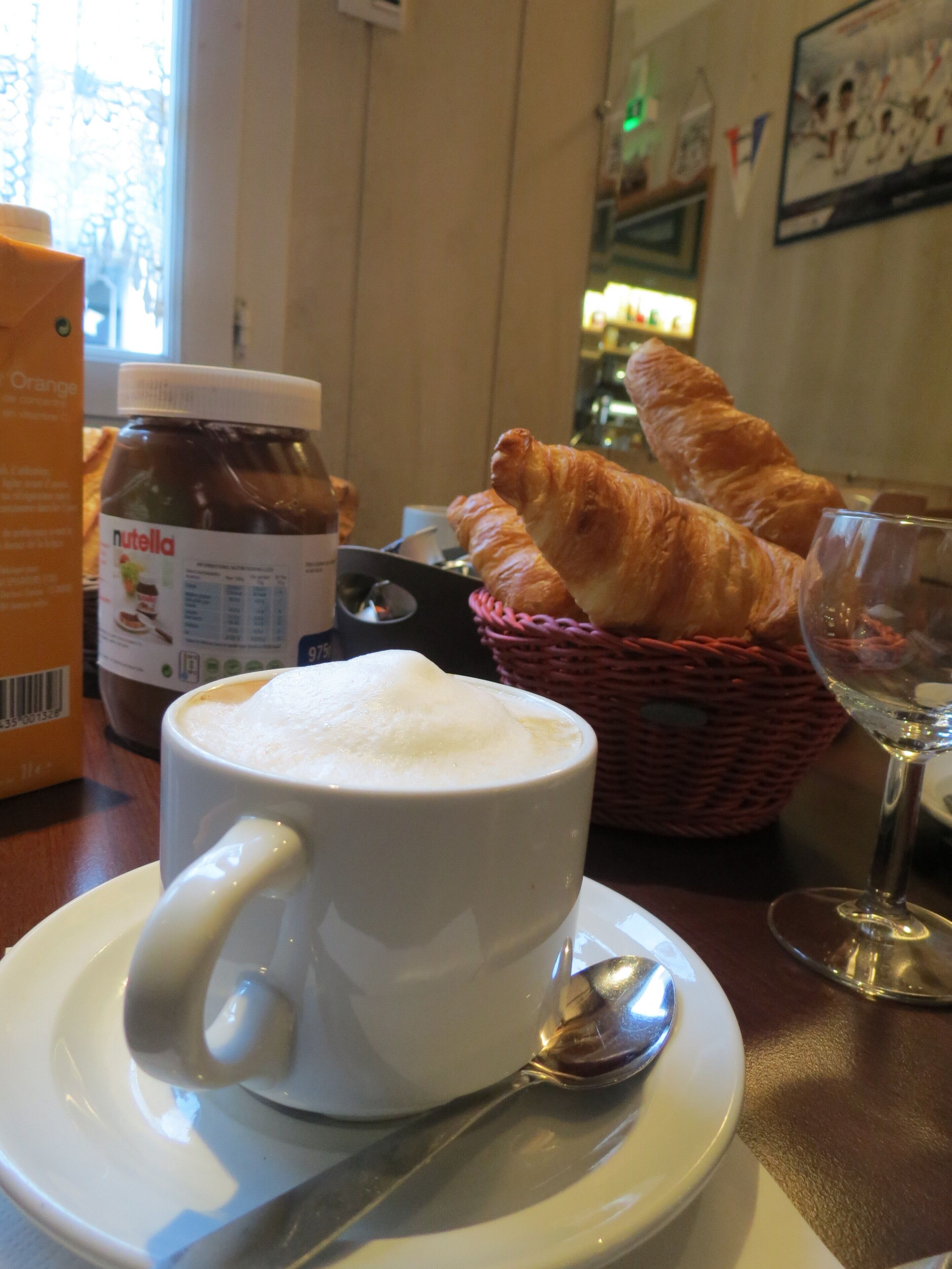 A cup of coffee with foam on top, a jar of Nutella, croissants, a basket, and a wine glass on a table.