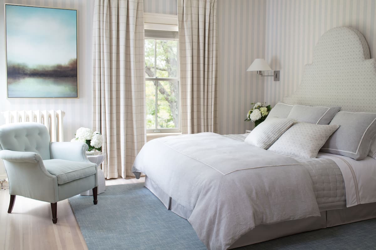 A bedroom with a large bed, a chair, a window with curtains, and a wall-mounted lamp.