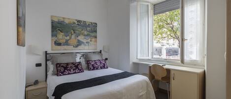 A bedroom with a bed, a desk, a chair, a window, and a painting on the wall.