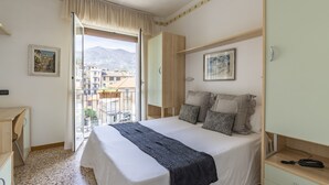 A bedroom with a bed, a wardrobe, a desk, and a window with a view of buildings and mountains. - Hotel Aida (Alassio)