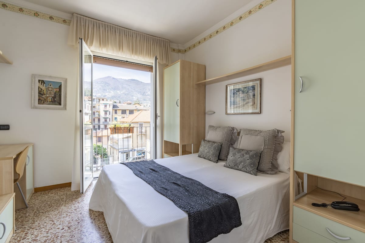 A bedroom with a bed, a wardrobe, a desk, and a window with a view of buildings and mountains.