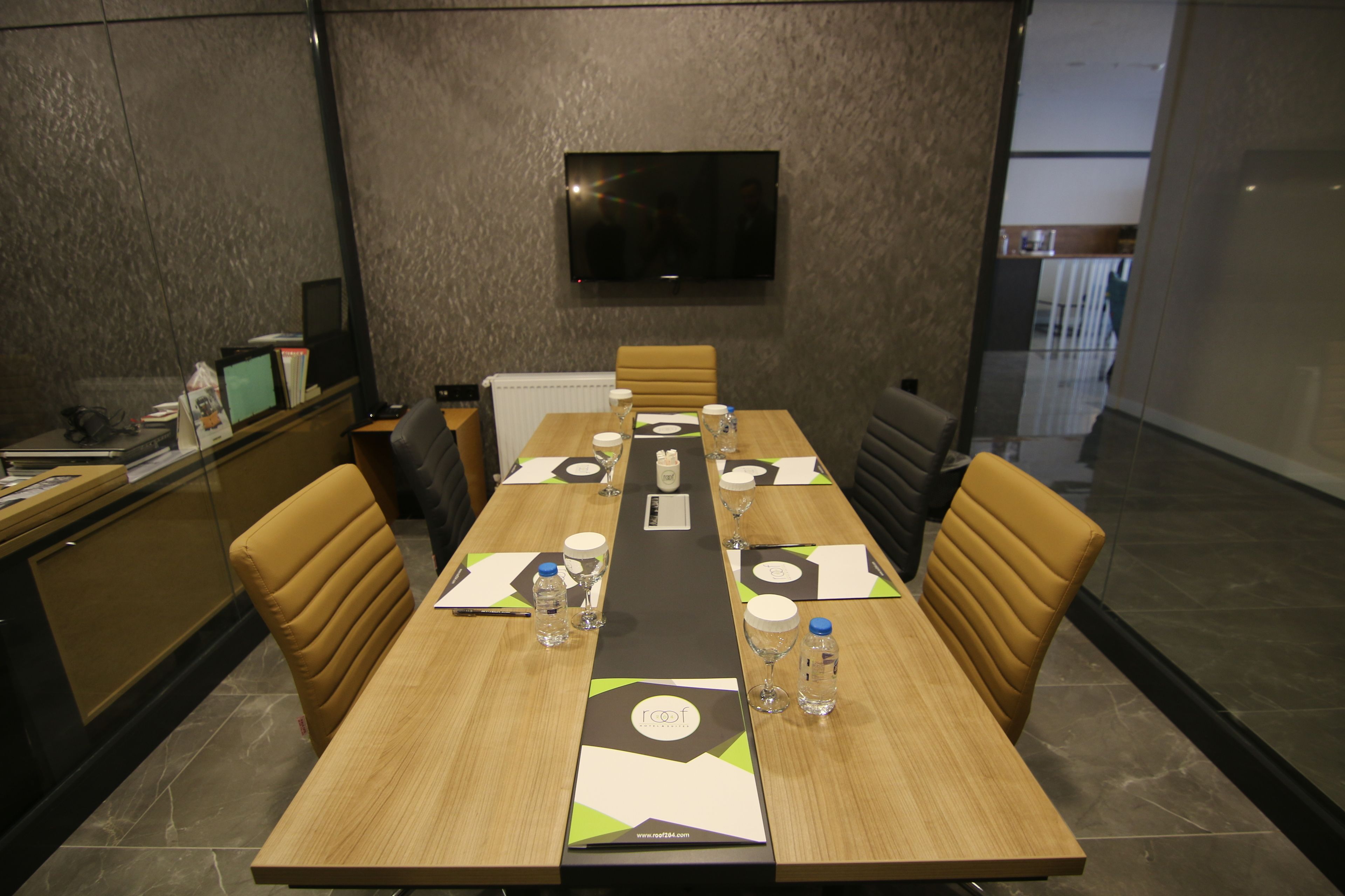 A conference room with a long table, chairs, a TV mounted on the wall, and a glass partition.
