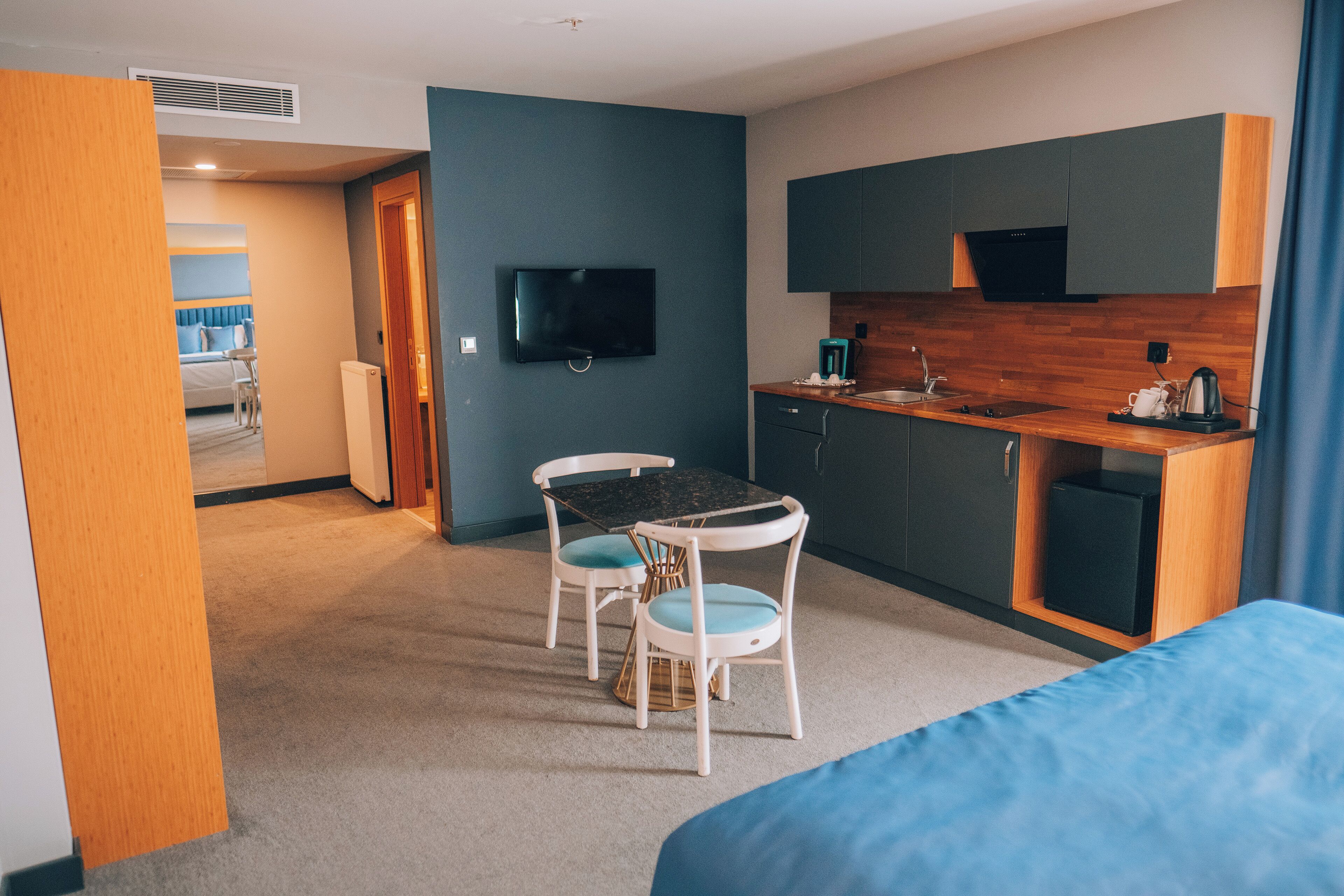 A modern hotel room with a kitchenette, a dining area with a table and chairs, a bed, and a TV mounted on the wall.