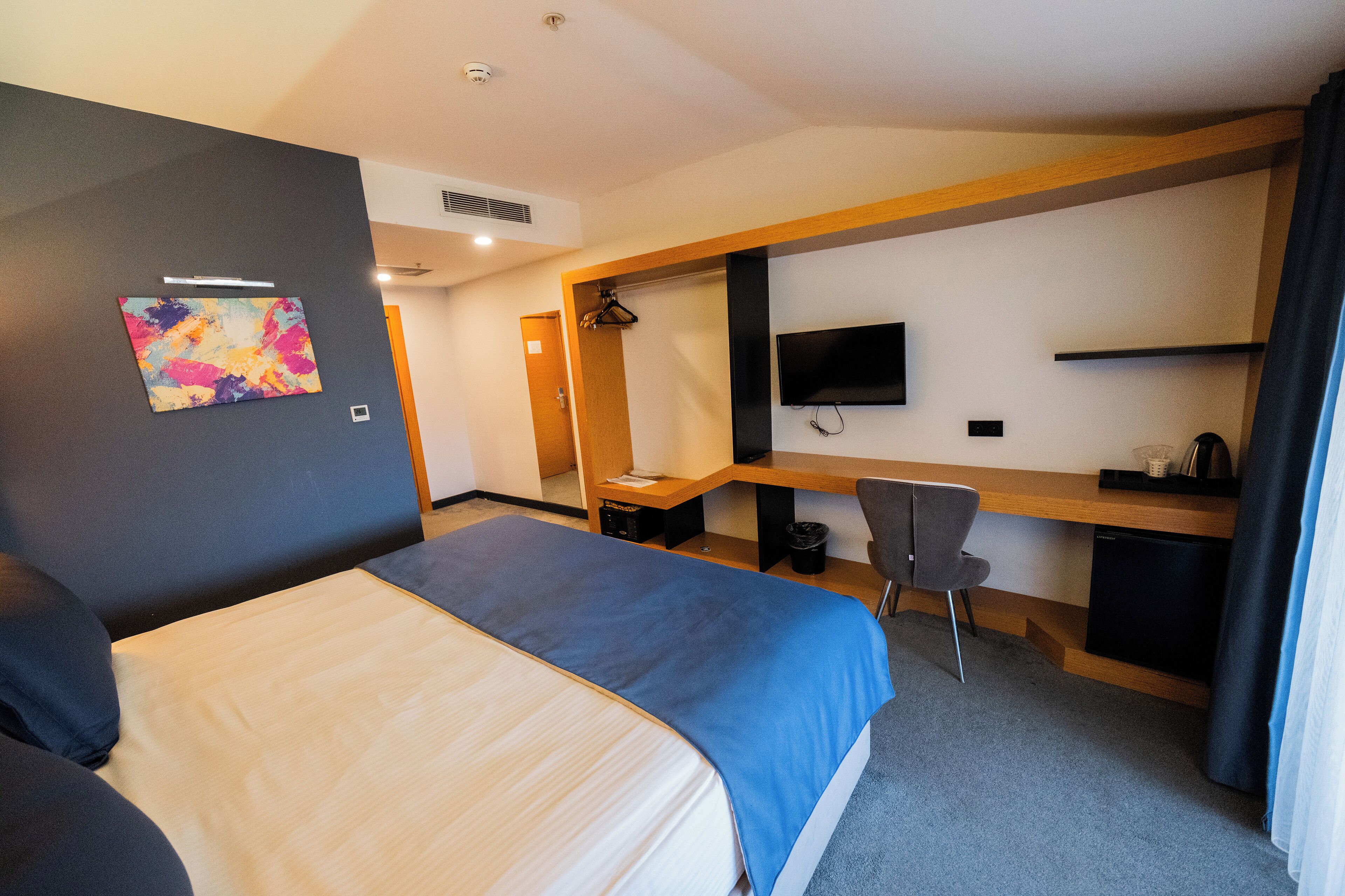 A hotel room with a bed, desk, chair, TV, and artwork on the wall.
