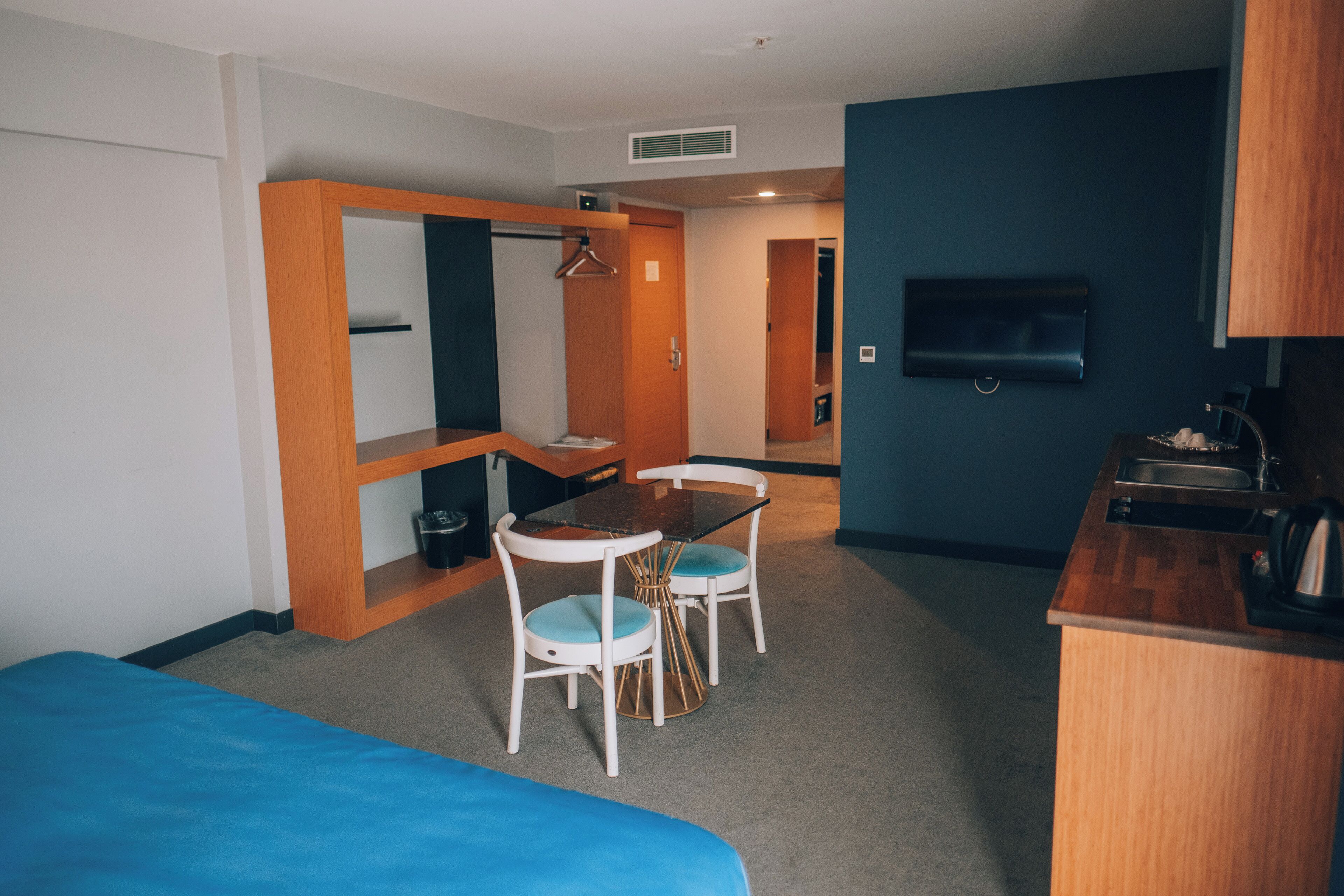 A compact hotel room with a bed, a small dining table with chairs, a kitchenette, a TV mounted on the wall, and a wooden wardrobe.