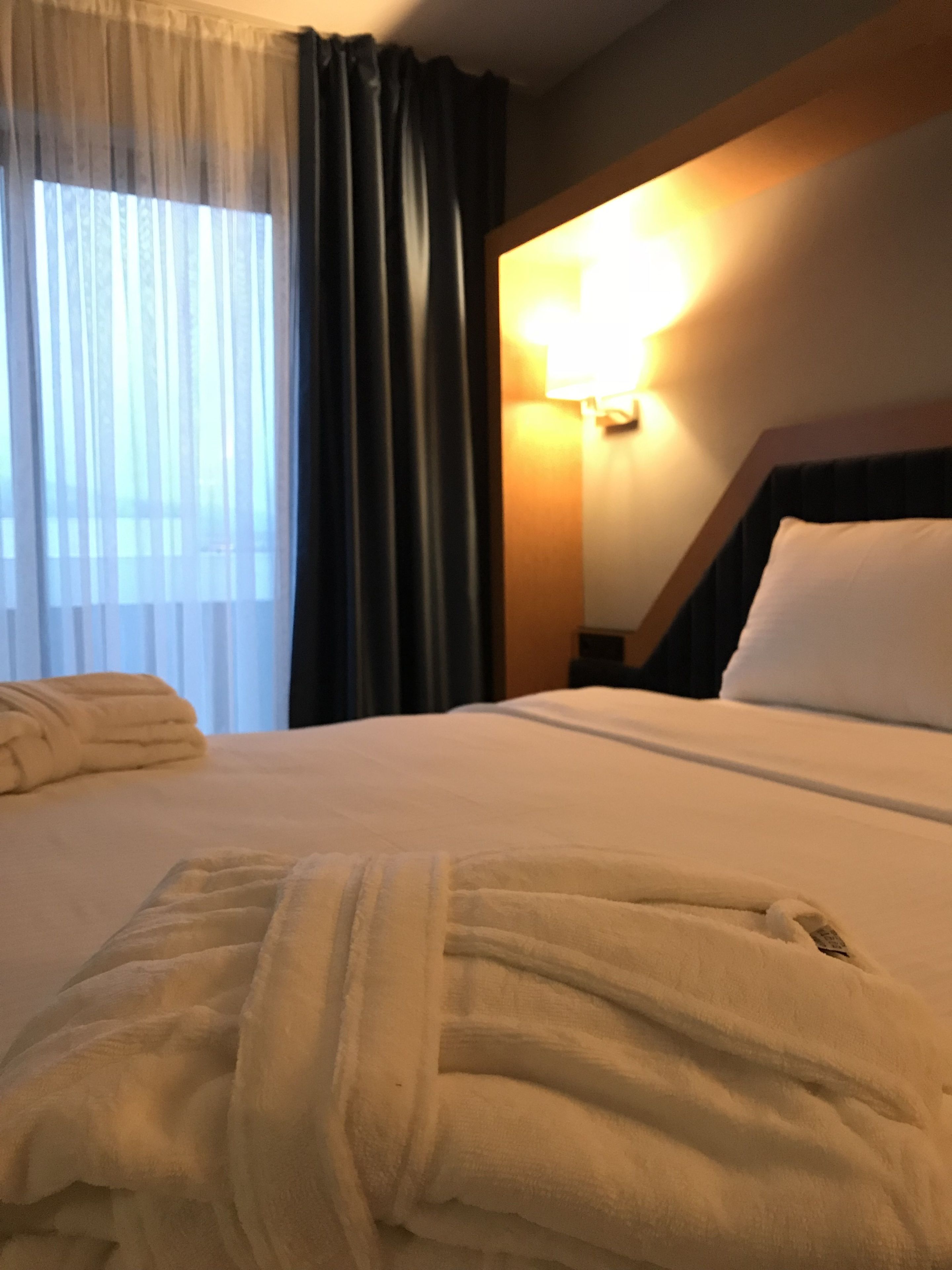 Una habitación de hotel con una cama bien hecha, cabecera con lámpara y ventana con cortinas.
