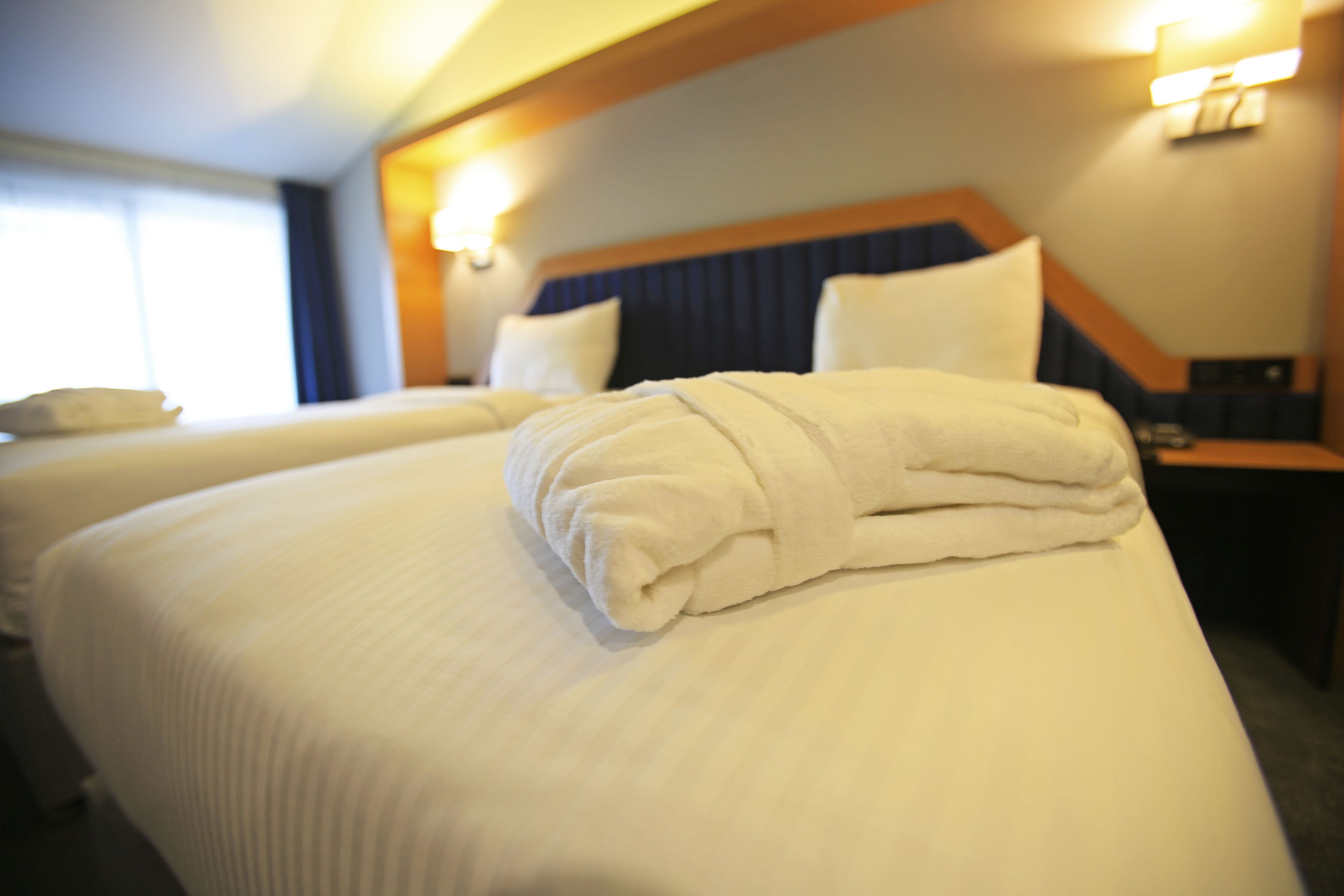 Ein Hotelzimmer mit zwei Betten, jeweils mit weißer Bettwäsche und einem gefalteten Handtuch. An der Wand sind Lampen angebracht und es gibt ein Fenster mit Vorhängen.