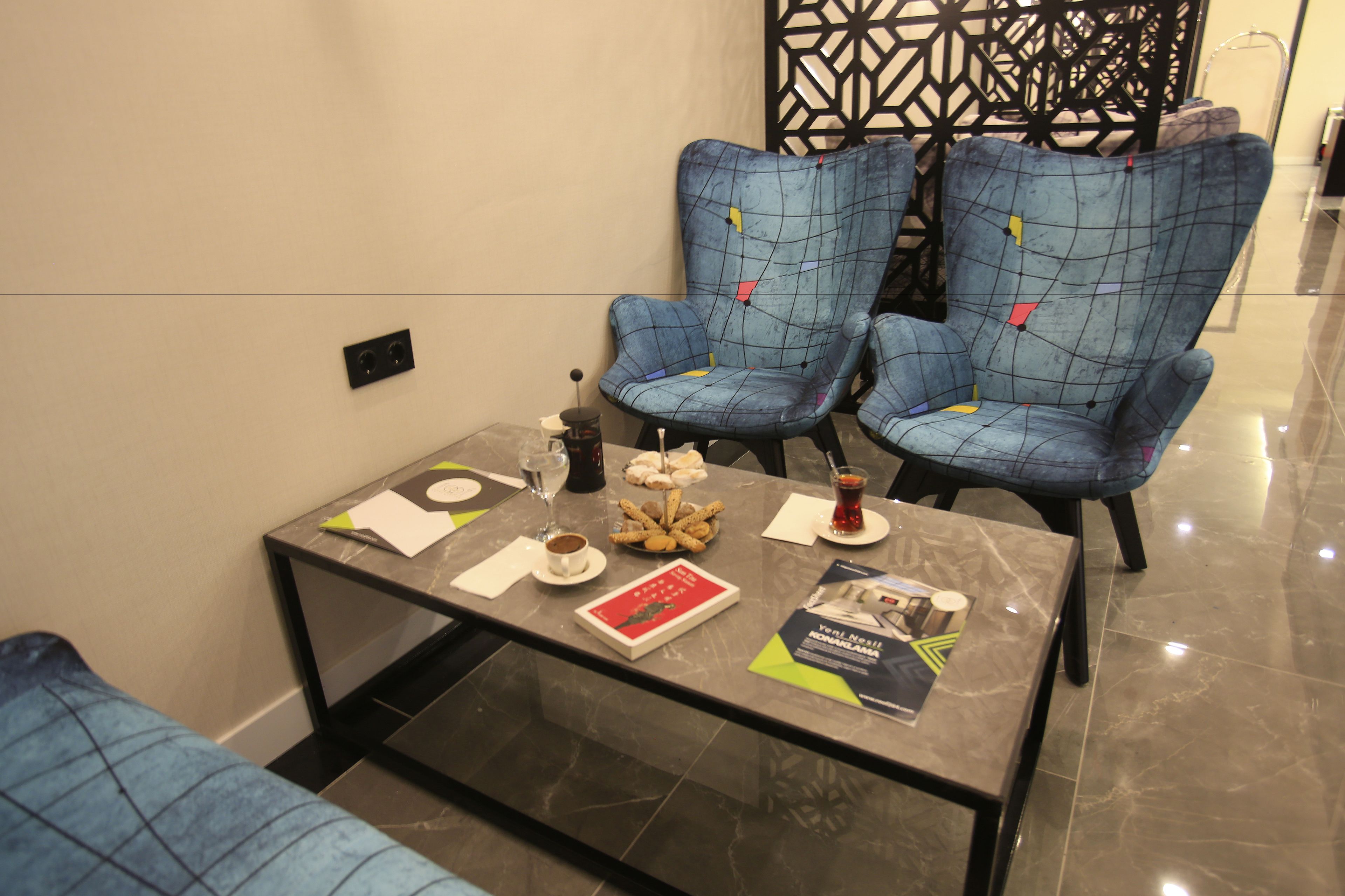 A modern hotel room with a glass-top table, two blue patterned chairs, a decorative wall panel, and a small selection of snacks and drinks.