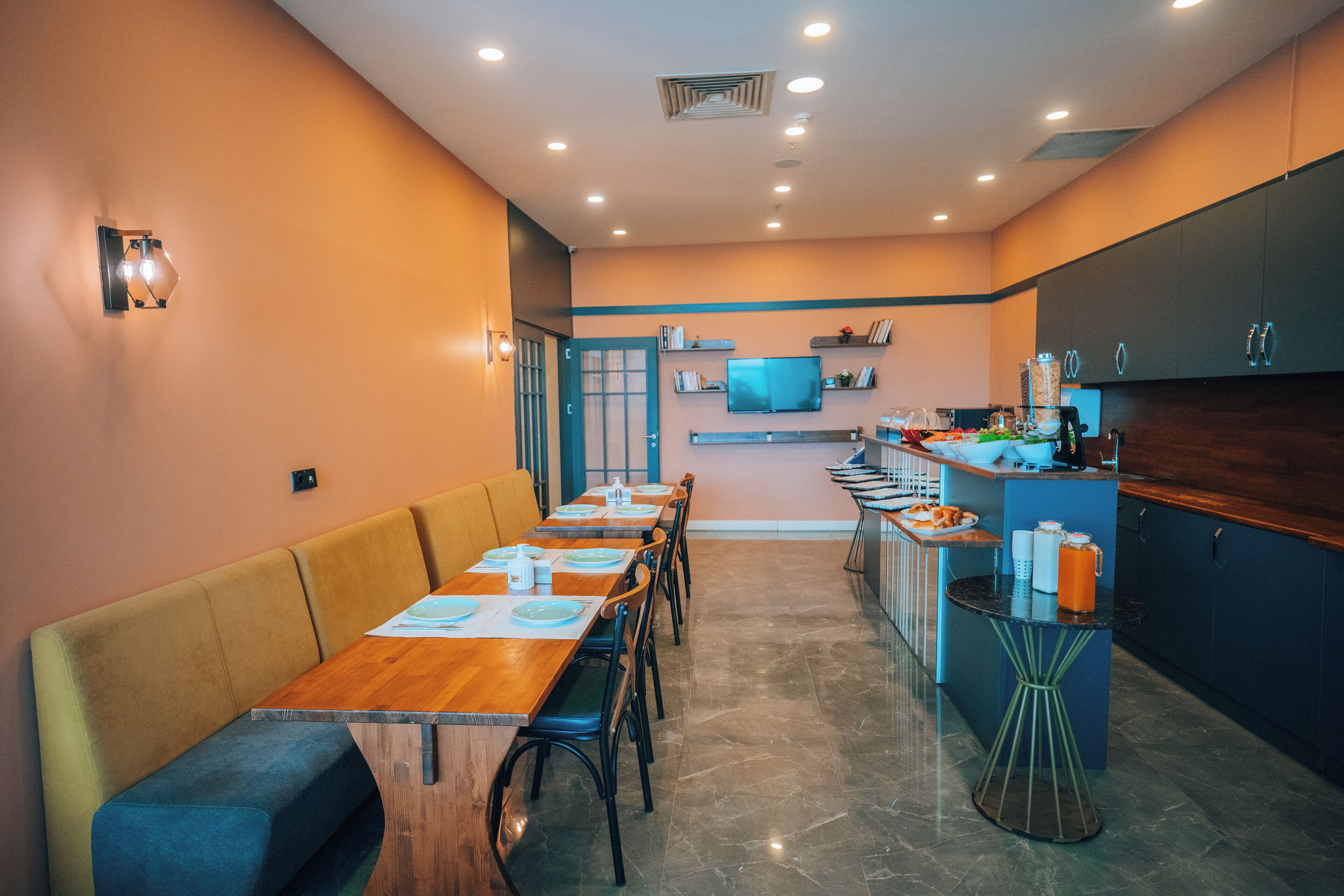 Un restaurant moderne avec une longue table en bois et des sièges rembourrés, un coin cuisine équipé d’un téléviseur et un petit comptoir de bar.