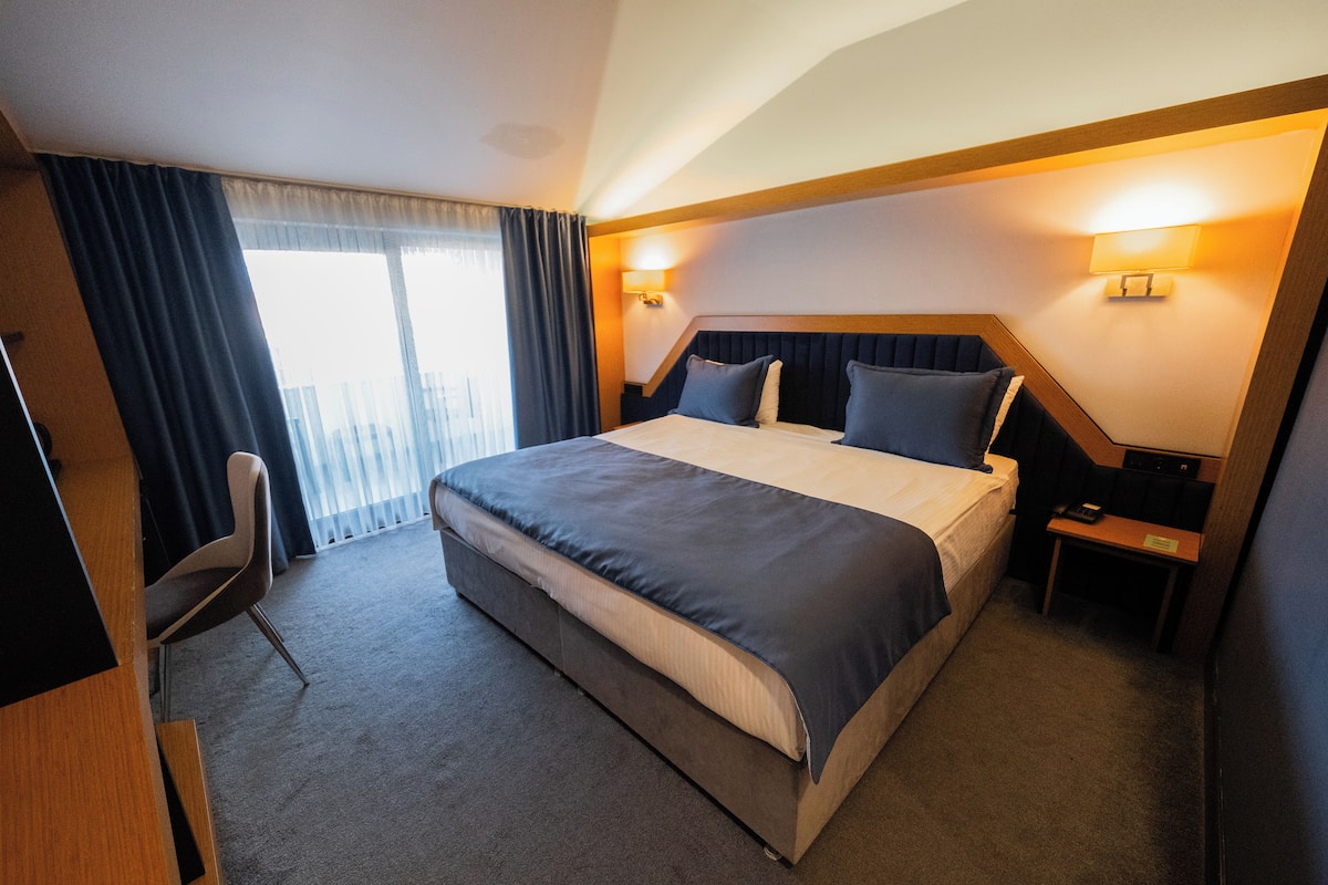 Une chambre d’hôtel avec un grand lit, un bureau et une chaise. Il y a deux lampes fixées au mur et une fenêtre avec des rideaux.
