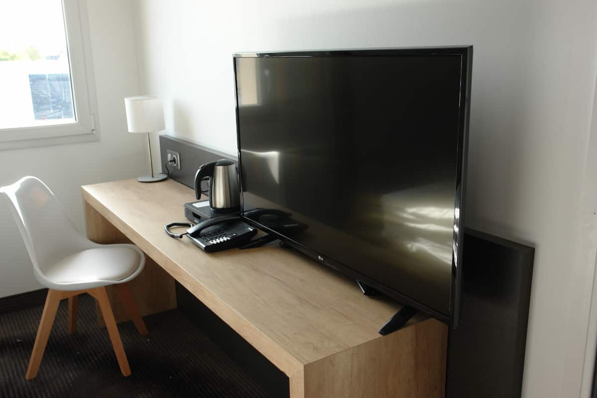 A hotel room with a wooden desk, a flat-screen TV, a white chair, a lamp, and a coffee maker.