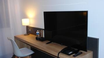 Een hotelkamer met een houten bureau, een flatscreen-tv, een lamp, een waterkoker, een waterfles, een stoel en een telefoon.