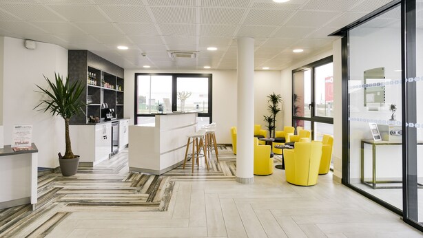 A modern reception area with a white reception desk, yellow seating, and a large window offering a view of the city. - Brit Hotel Confort Loches (Loches)