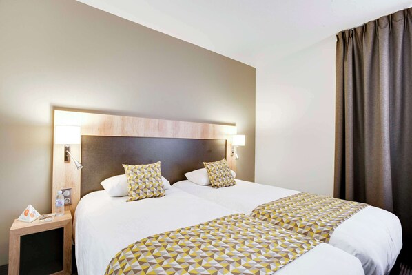 A hotel room with two beds, a wooden headboard, bedside lamps, and a nightstand with a water bottle and a brochure. - Brit Hotel Confort Loches (Loches)