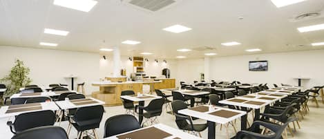 A conference room with rows of tables and chairs, a kitchenette, and a large plant.