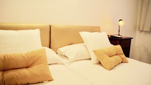 Una cama bien hecha con almohadas blancas y amarillas, una mesita de noche de madera con una lámpara y una pared lisa.
