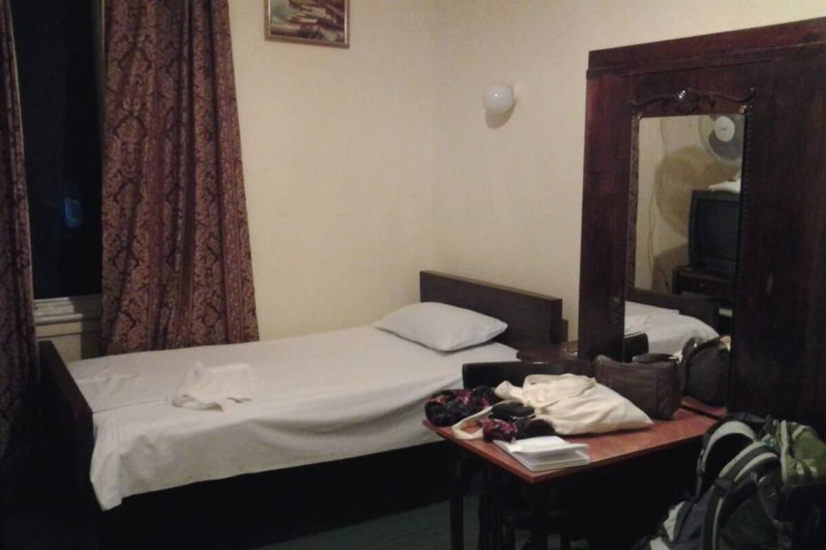 Una habitación pequeña y poco iluminada con una cama individual, un escritorio de madera y un espejo. Hay maletas y un almohadón sobre el escritorio.