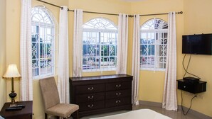 Habitación de hotel con cama, cómoda, silla, televisor montado en la pared y tres ventanas con cortinas.