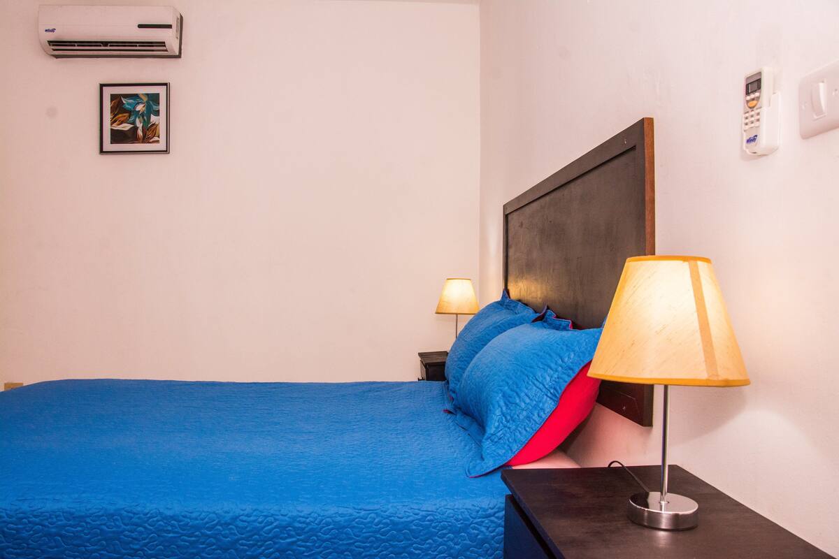 A hotel room with a bed, two bedside lamps, a wall-mounted air conditioner, a framed picture, and a wall-mounted telephone.