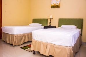 A hotel room with two beds, beige bedspreads, a green headboard, a nightstand with a lamp, and a floral painting on the wall.