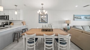 A modern kitchen with white cabinetry, a wooden dining table, and a chandelier. There is a sofa and a lamp in the living area. - Seacrest - 6 - 2 Br townhouse by RedAwning (Panama City Beach)