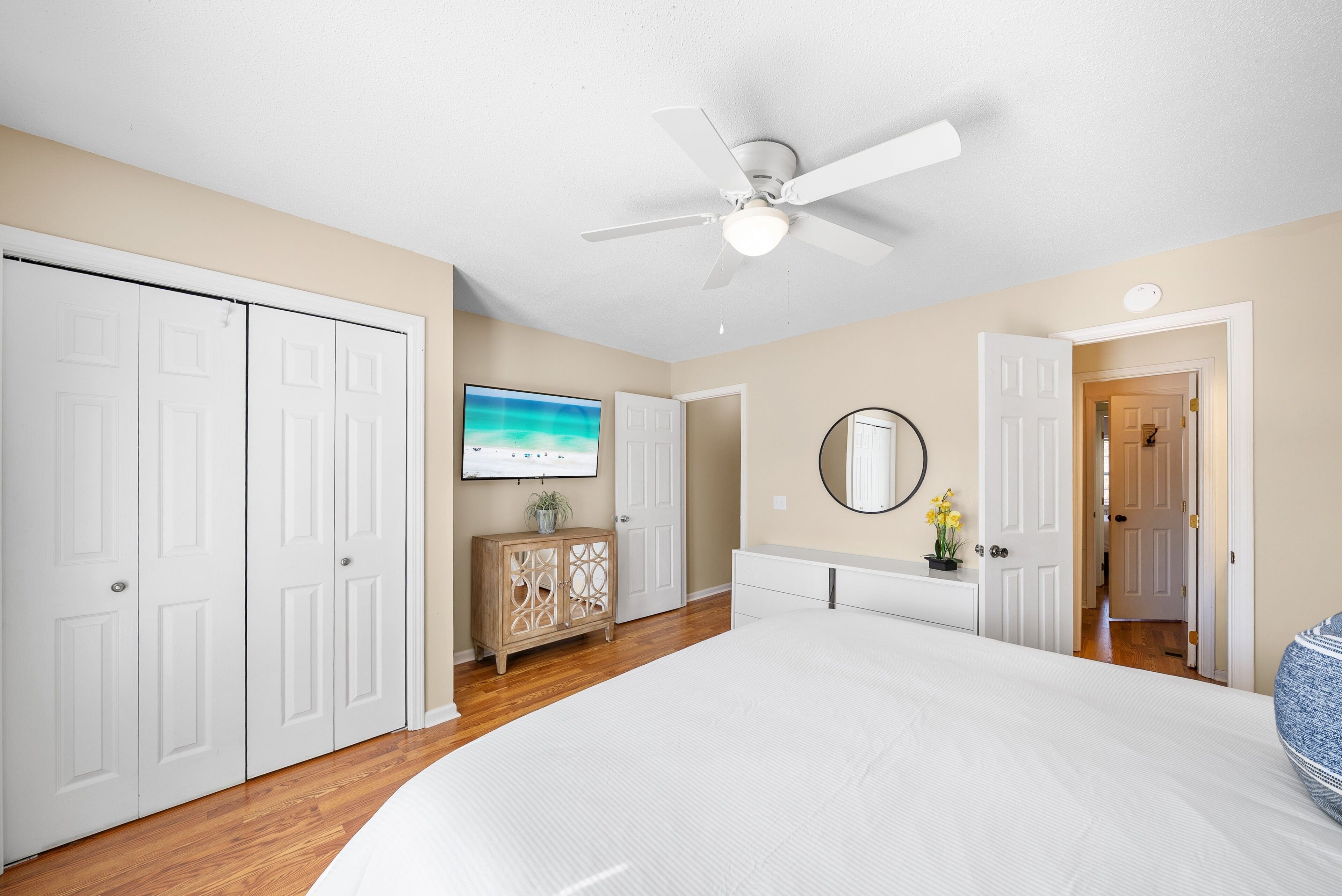 A bedroom with a large bed, a ceiling fan, a TV, a dresser, and a door leading to another room.