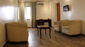 Una habitación de hotel con piso de madera, un escritorio con una silla, dos sillones, un televisor montado en la pared y un equipo de aire acondicionado.