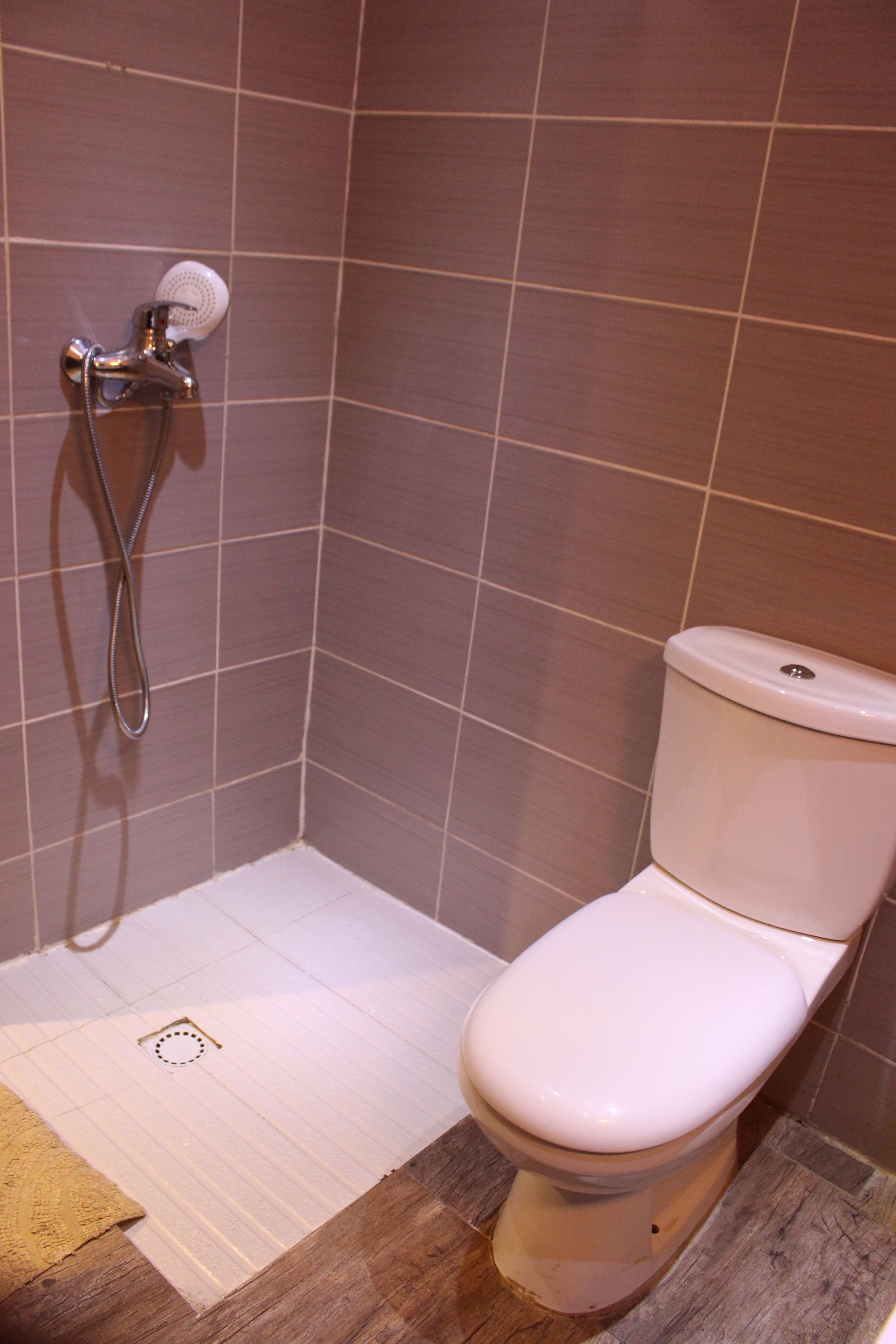 Un baño con inodoro blanco, ducha con cabezal de mano y paredes de azulejos marrones.