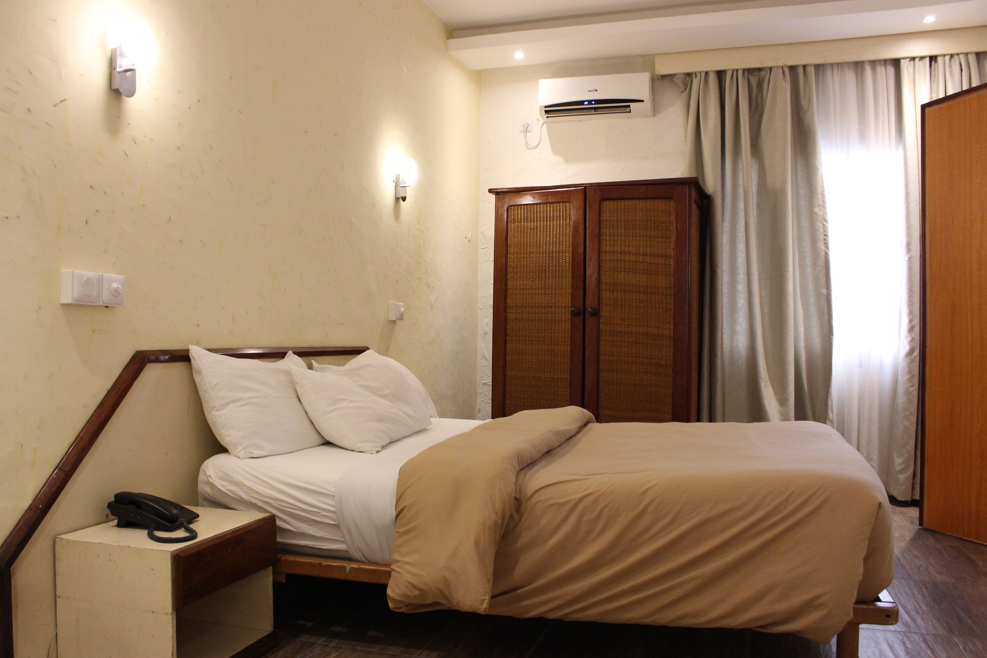 A hotel room with a bed, a bedside telephone, an air conditioning unit, and a wardrobe.