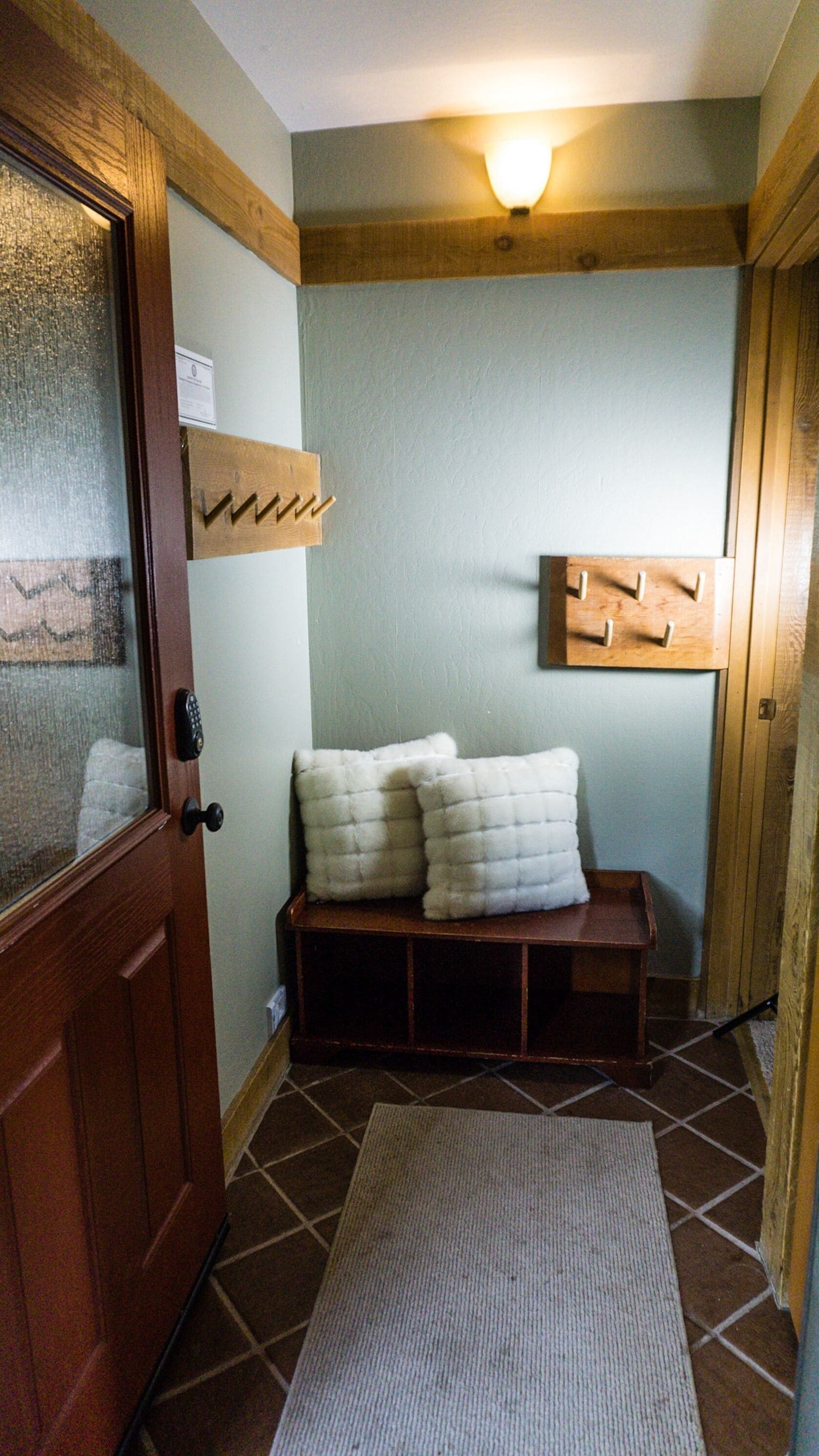 A small, cozy room with a wooden door, a bench with cushions, a wall-mounted coat rack, and a light fixture.