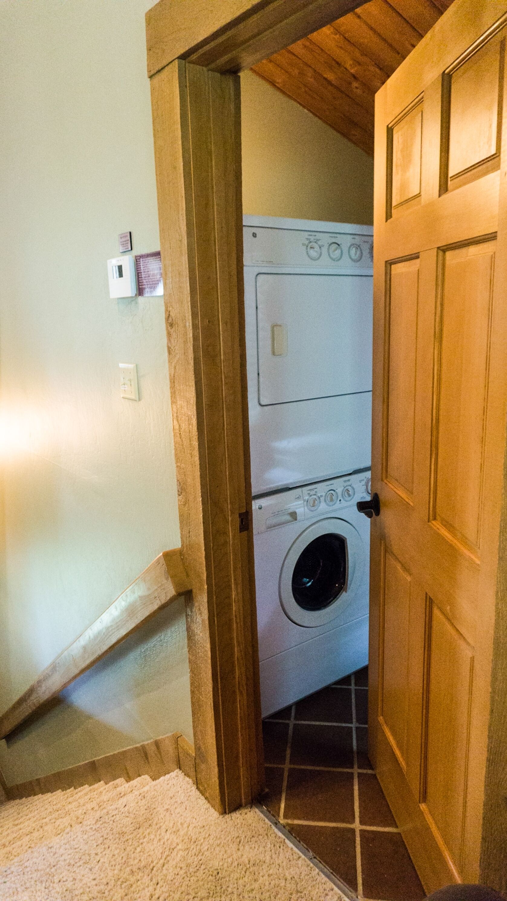 A laundry room with a stacked washer and dryer, wooden door, and a small step leading up to the room.