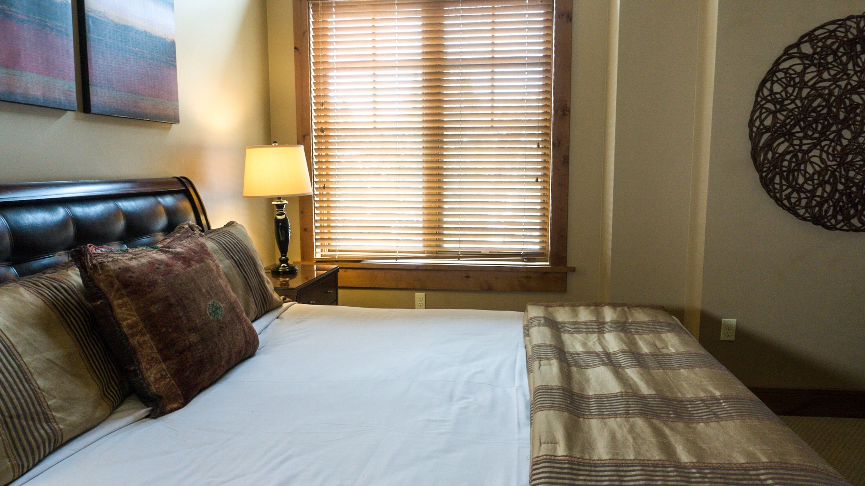 A hotel room with a bed, a nightstand, a lamp, a window with blinds, and a wall decoration.