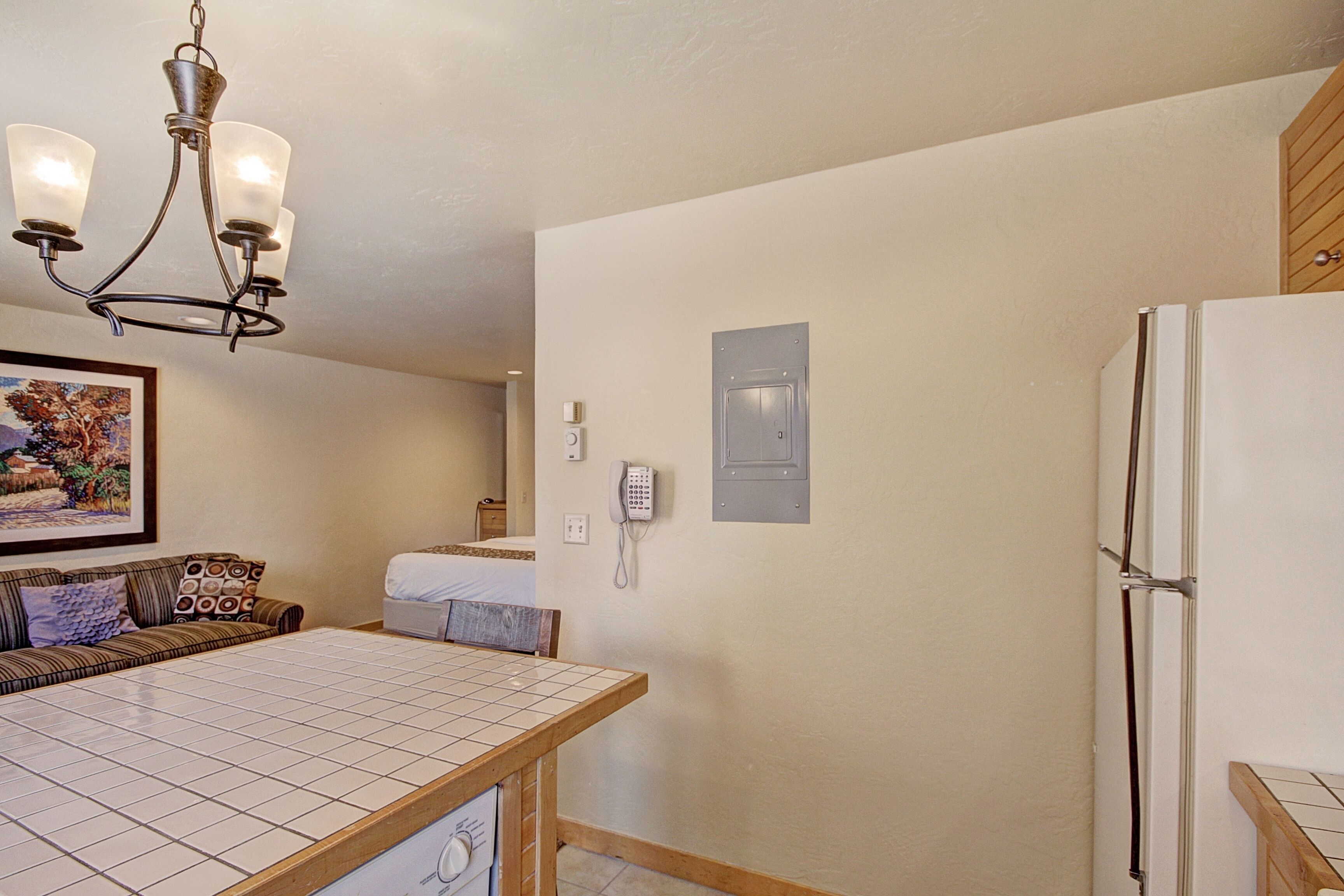 A kitchen with a tiled countertop, a refrigerator, a phone, and a wall-mounted light fixture.