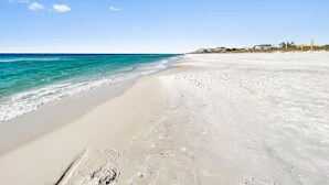 A beach with clear turquoise water, white sand, and houses in the background.