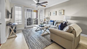 A living room with a beige sofa, a coffee table, and a ceiling fan. There is a door leading to a patio area with a chair and a table.