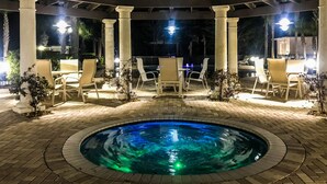 An outdoor pool area with a circular pool, surrounded by white chairs and tables, under a covered pavilion with columns.