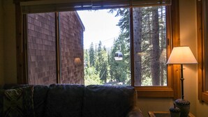 A room with a large window, a sofa, a lamp, and a view of a forest and a cable car. - Ski Trails 4024 (Truckee)